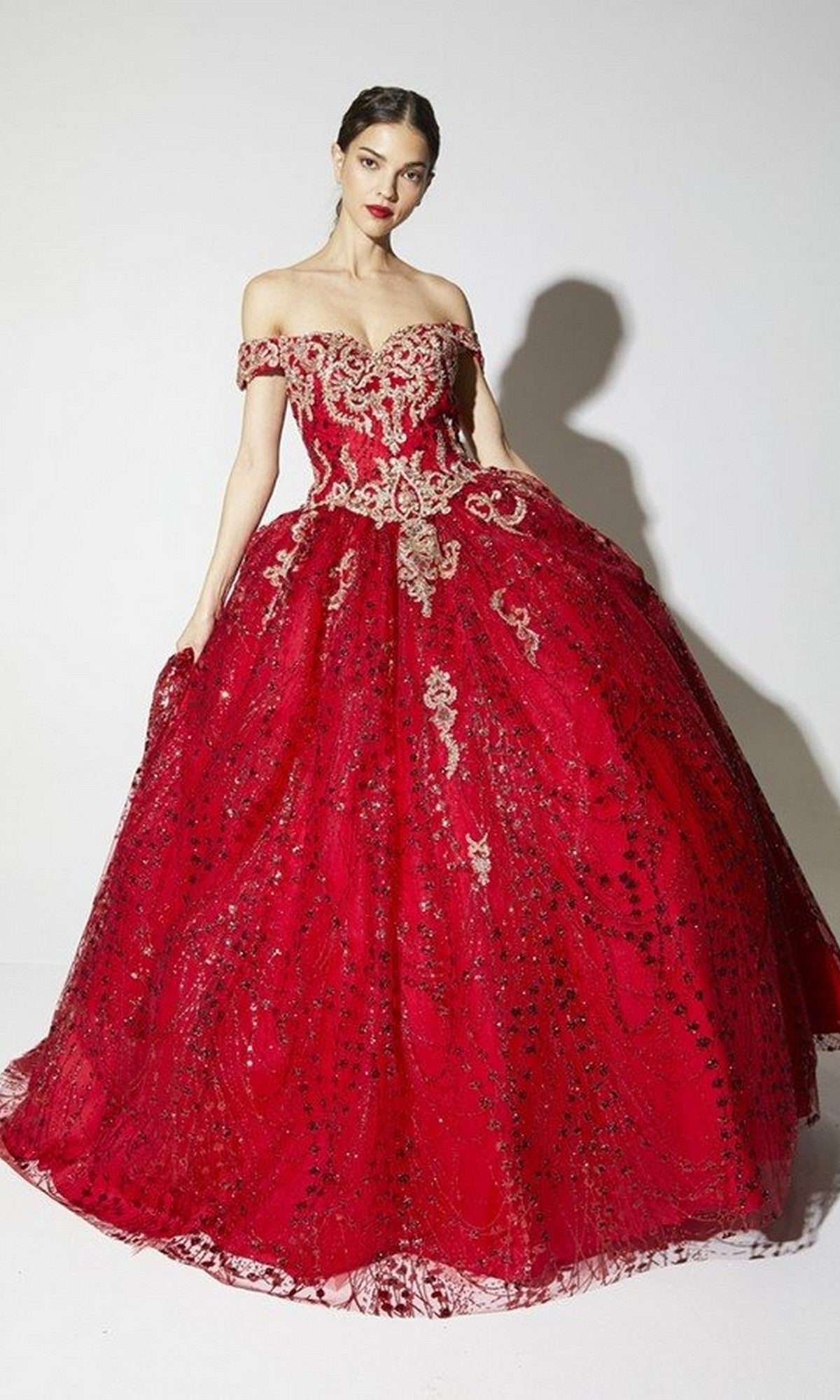 A woman wearing the Eureka 9778 long red prom dress with an off-the-shoulder neckline, glitter fabric, gold embroidery, and sequin details stands against a plain white background.